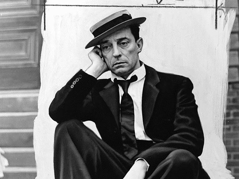 Buster Keater sitzend - den Arm auf das rechte Knie und den Kopf auf die rechte Hand gestützt, schaut er vor sich hin.